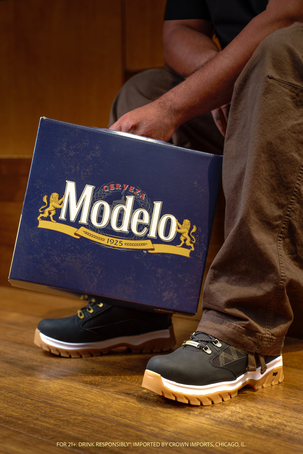 A person sitting on a wooden floor holds a large blue Modelo shoe box while wearing the Wilderness boot from the collection. 